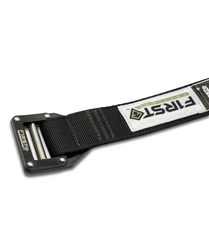 Backside shot of then black nylon tactical belt with a black metal buckle, featuring a white brand patch with 'FIRST' and a green diamond logo, shown on a white background |color_black